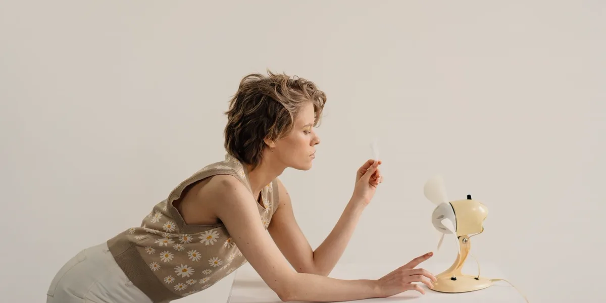 A person leaning over a table towards a small desk fan